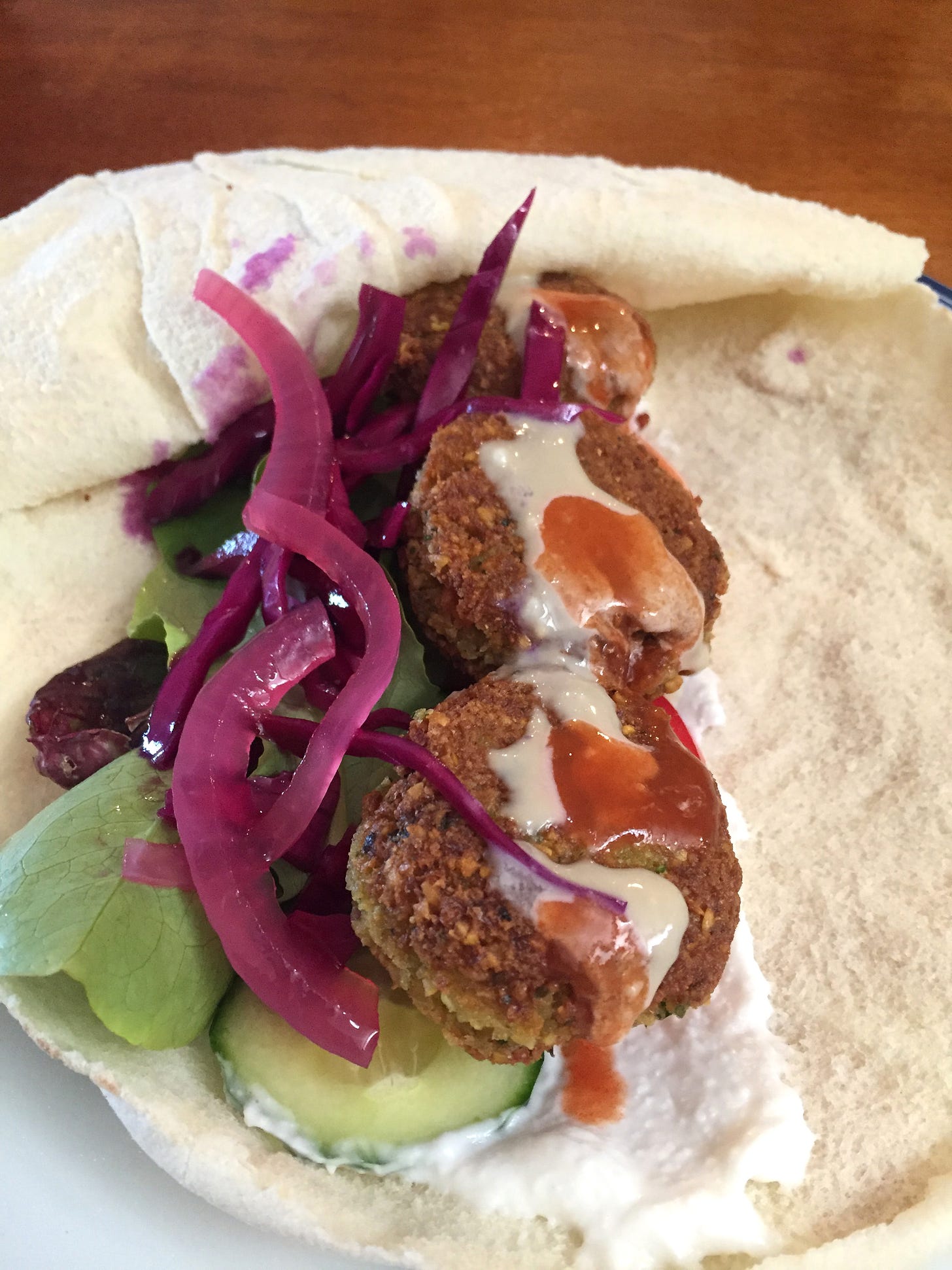 an opened pita pocket filled with falafel patties, cucumber, lettuce, and pickled red cabbage and onion. White garlic sauce is visible on the bottom, and a drizzle of tahini and hot sauce on top.