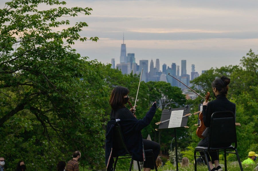 Two violinists are seen seated among greenery; the buildings of New York City are visible in the distance.