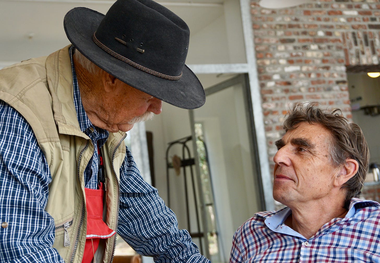 Two older men look at each other. One is wearing a hat, and they're both in plaid shirts.