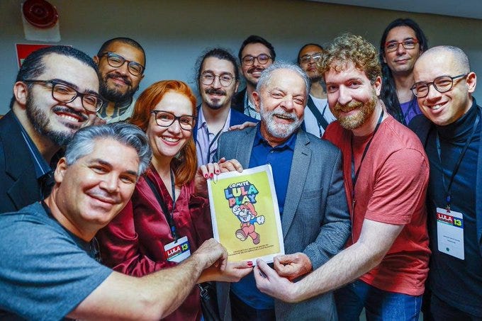 Lula aparece no centro da foto, segurando a cartilha Lula Play, que na traz um desenho do ex-presidente simulando um jogo de videogame. Junto com Lula, posam para a foto representantes do setor de games.