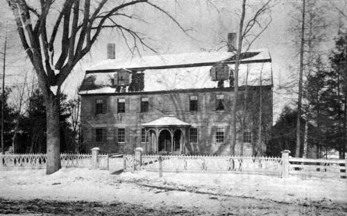 black and white photo of stone house with mansard roof and round front porch. House stands behind white fence and snow-covered lawn.   