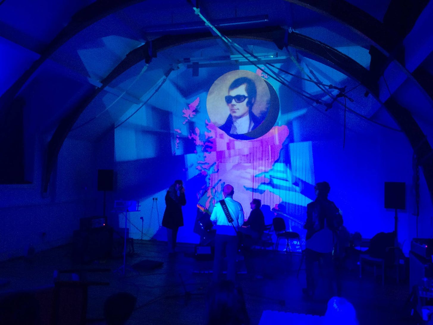 A large space with domed ceiling. An image of Robbie Burns is projected onto a map of Scotland onto the back wall. The lighting is blue and red.