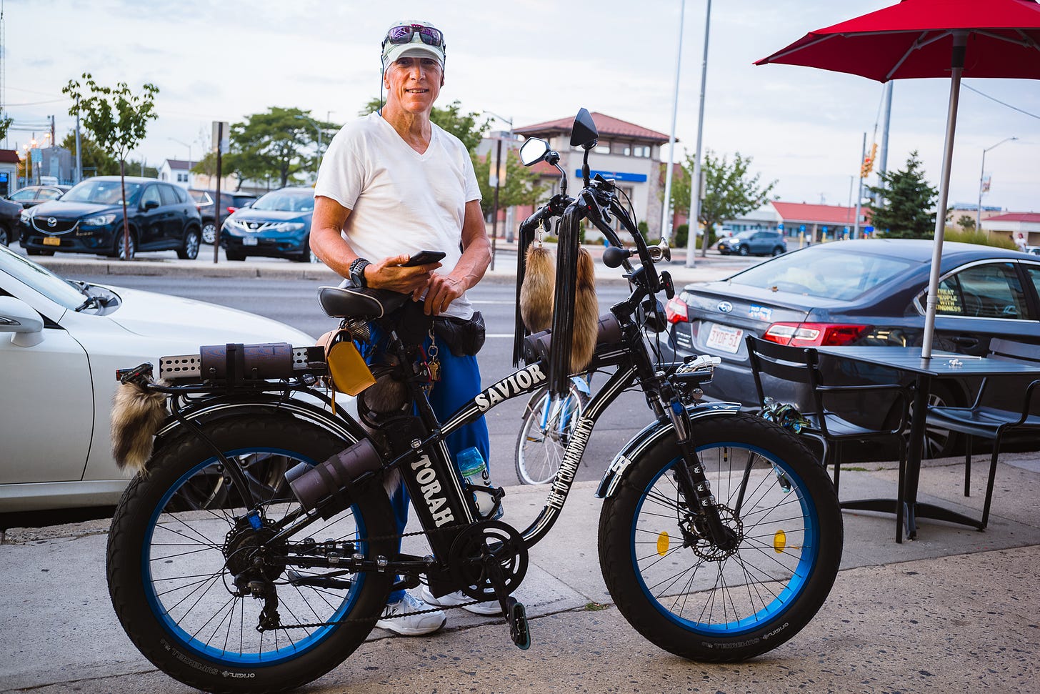 Man in a white cap with sunglasses pulled up on the peak, white v-neck t-shirt and blue pants behind his tricked-out black bicycle with furs and tassles hanging from the handlbars and 'TORAH' 'SAVIOR' and 'THE 10 COMMANDMENTS' written in white on the frame