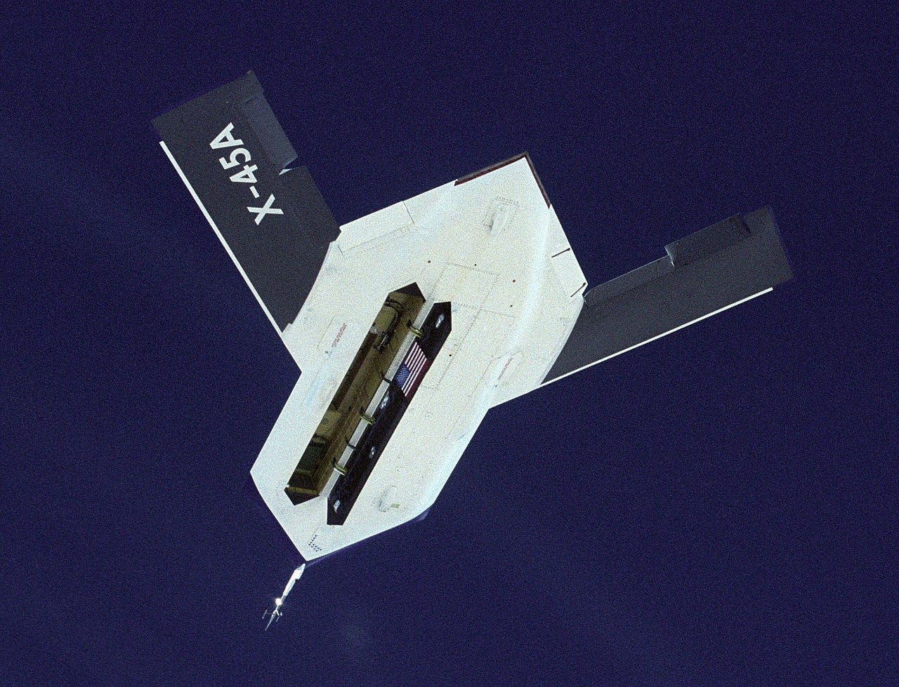 A sharply angular drone in flight against a grainy blue sky, with bomb bay open.