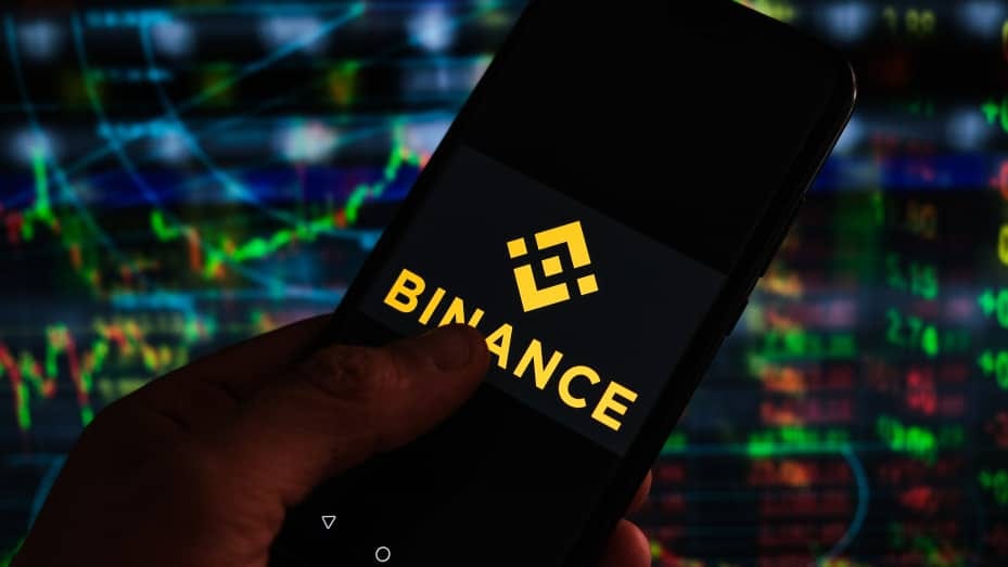 The logo of cryptocurrency exchange Binance displayed on a smartphone with stock market percentages in the background.
