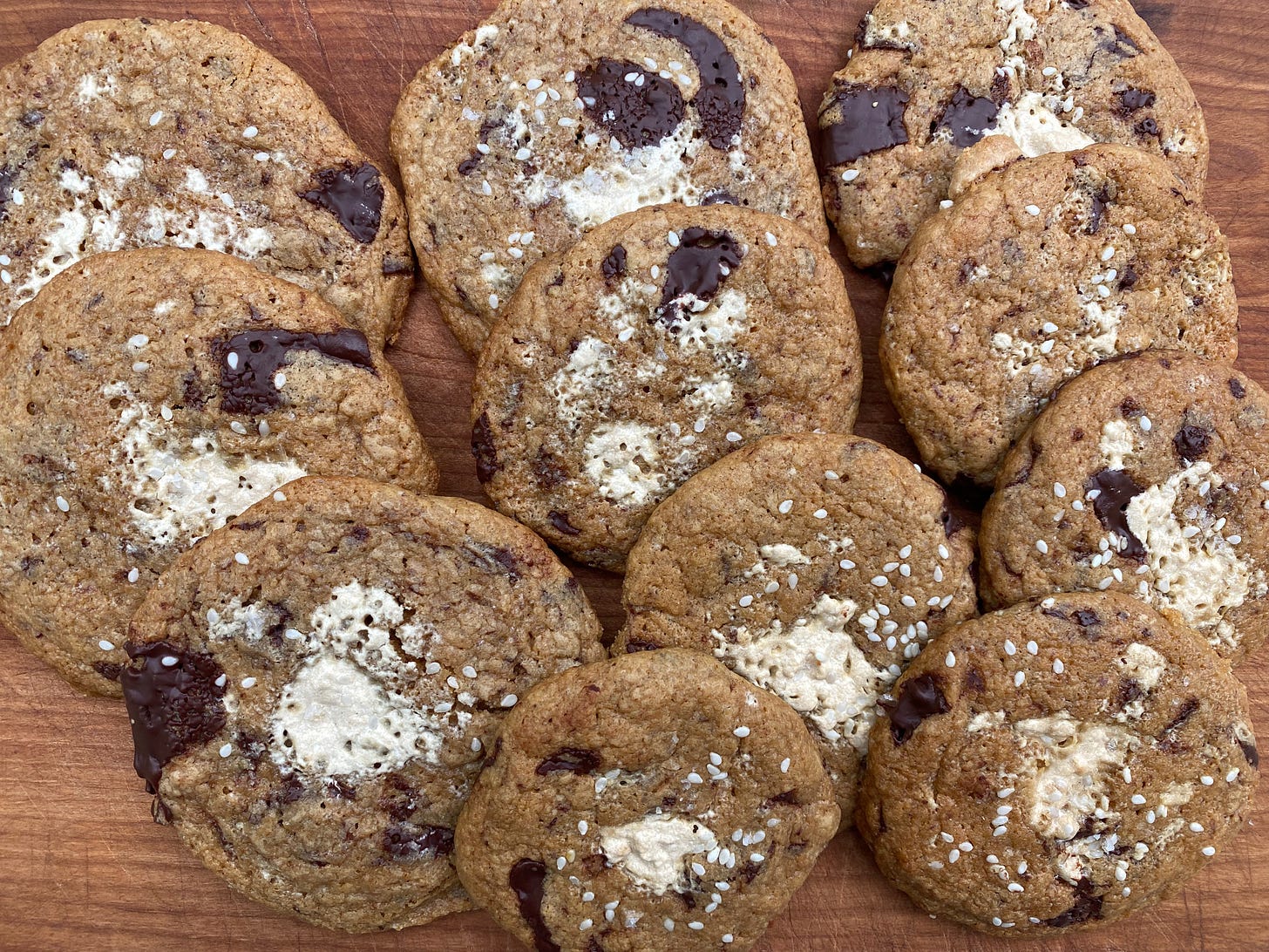 Many large cookies stacked in overlapping rows on a wooden cutting board. They all have melted pools of halvah and big chunks of chocolate, and they’re topped with sesame seeds.