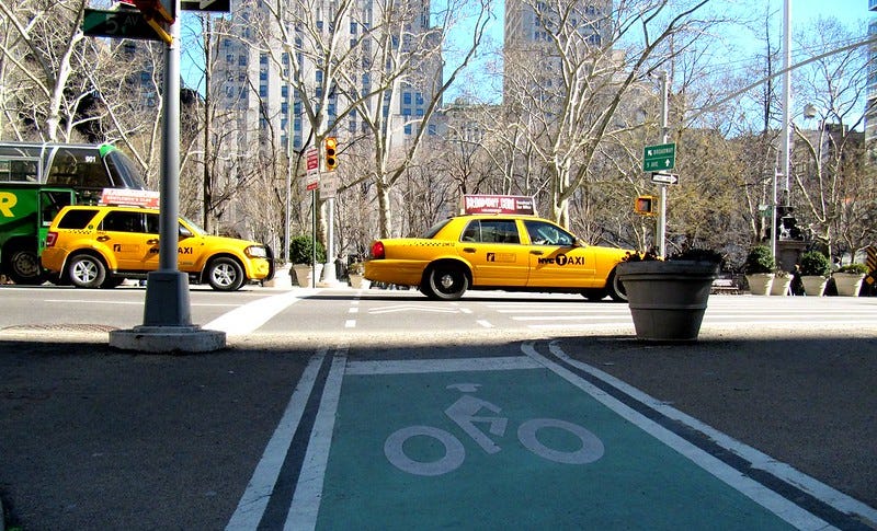 A protected bike lane in the foreground as New York City taxis pass in the background.