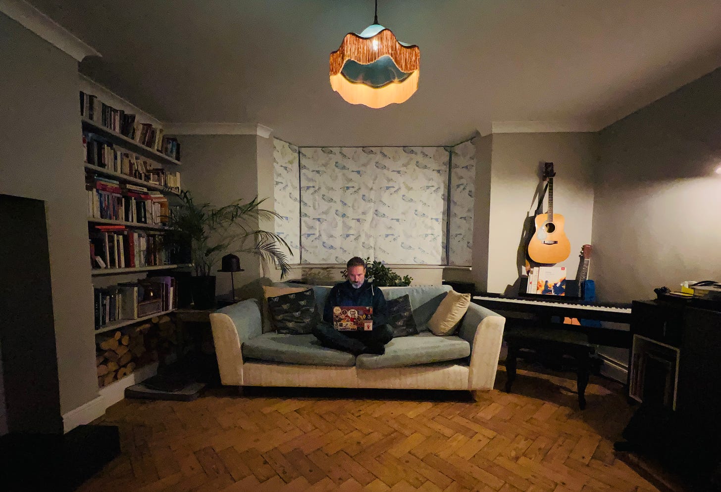 A wide angle photo of a writer in his living room. sat on a sofa with a laptop. A fire crackles out of shot and a guitar hangs on the wall behind an electric piano.
