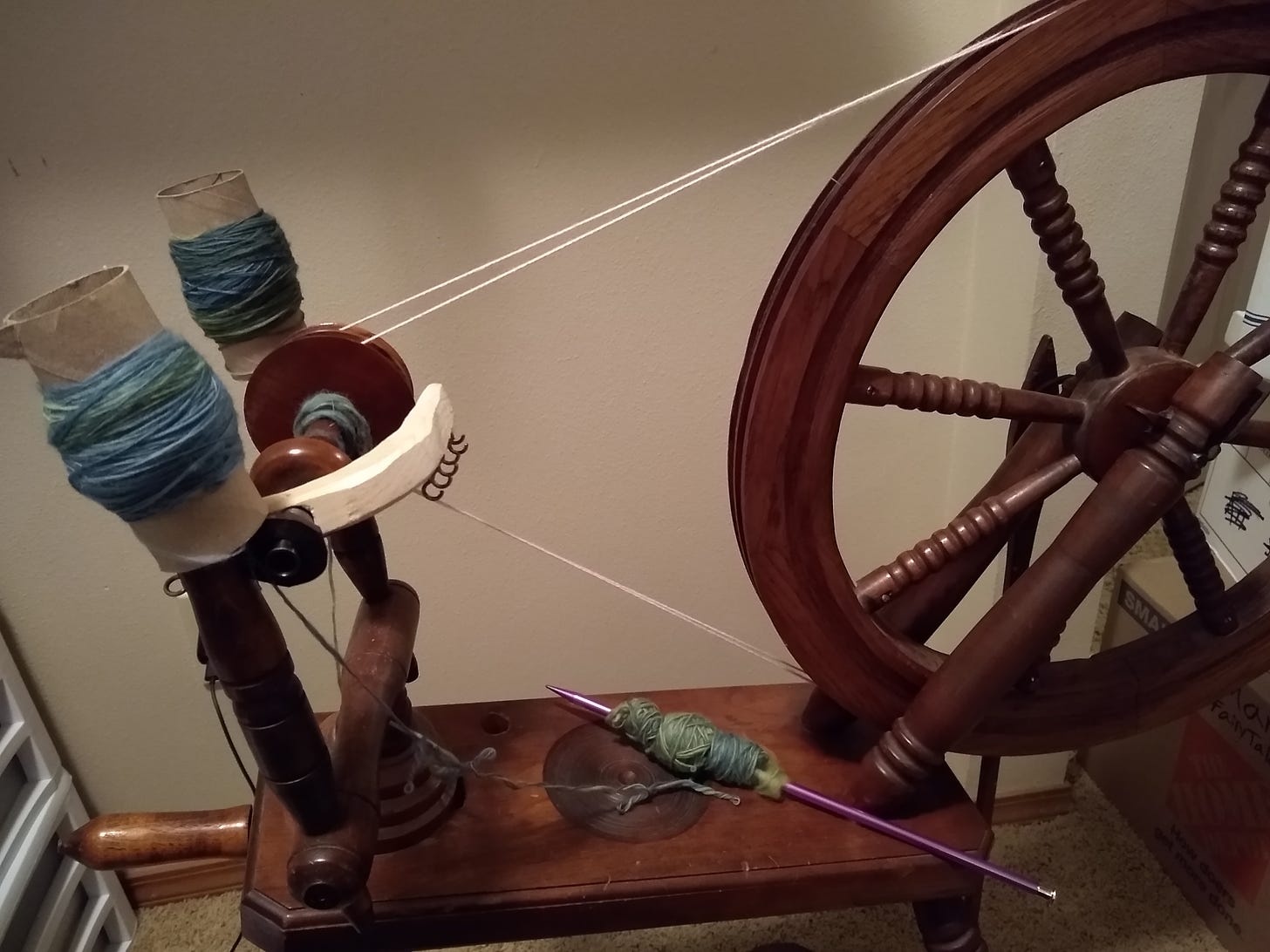 A spinning wheel with two toilet paper rolls of yarn on upright posts, and yarn wound onto a knitting needle