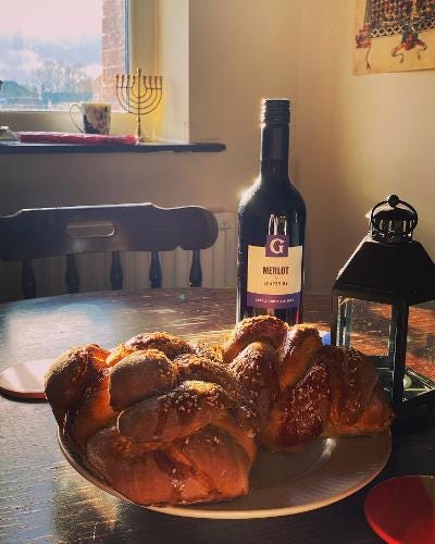 A photo of two loaves of challah and a bottle of wine on a wooden table, bathed in sunlight. The scenery of the room can be seen behind; a print of a medieval illustration of a menorah, and an actual menorah on the windowsill. A lantern sits near the bread on the table.