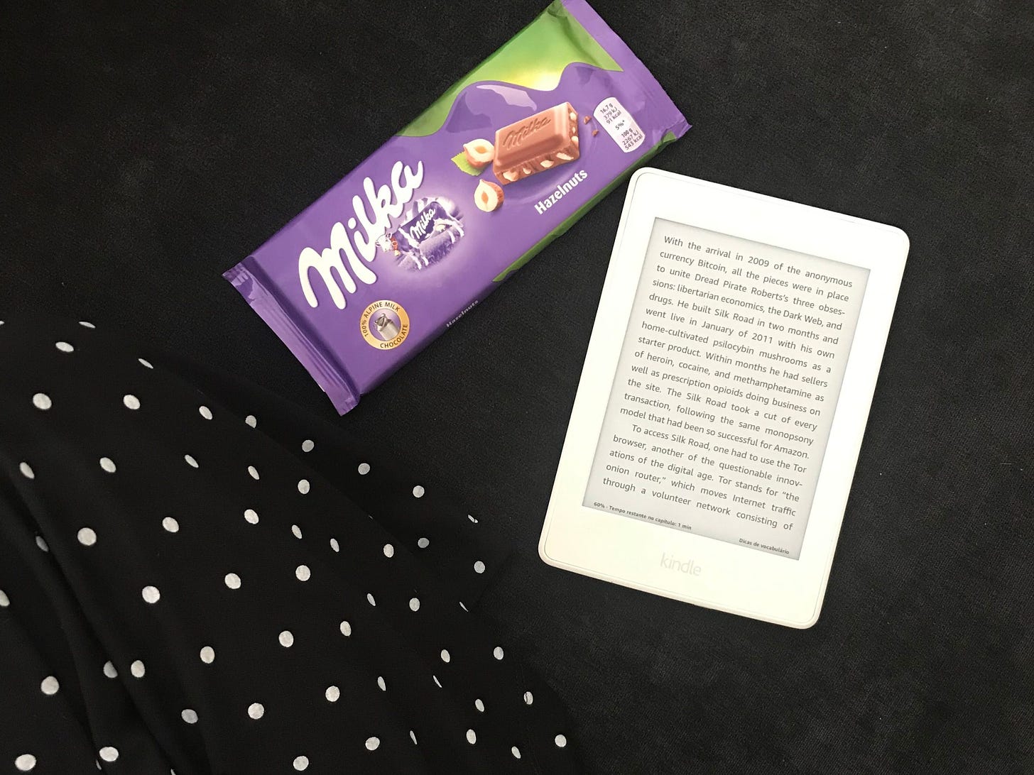 uma foto tirada de cima, de uma pessoa de pernas cruzadas com uma barra de chocolate milka e um kindle paperwhite branco mostrando uma página aleatória de um livro.