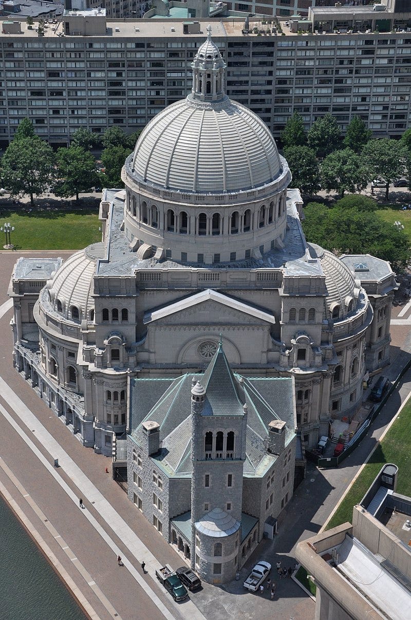 Aerial photograph of a triangular lot between roads and their sidewalks. The lot contains a small, Romanesque church filling the front point to the sidewalks, connected to a much larger and impressive domed, Neoclassical building behind it, filling the lot to the sidewalks to the left and right.