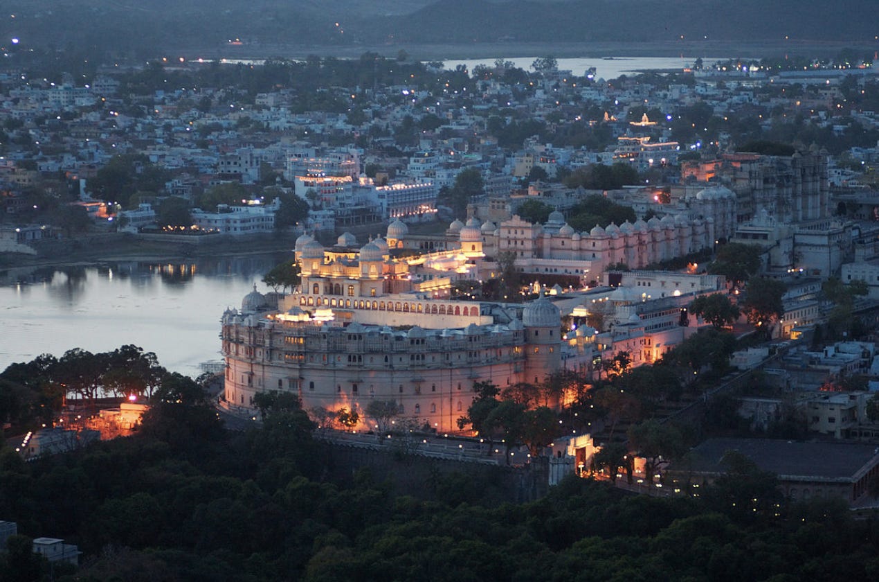 A city in the dusk light, showing many white buildings with many domes, interspersed with trees. Some of the buildings are lit up with golden lighting. In the middle left and in the central background, there are bodies of water reflecting the dusk light. The far background, there are mountains behind the body of water.