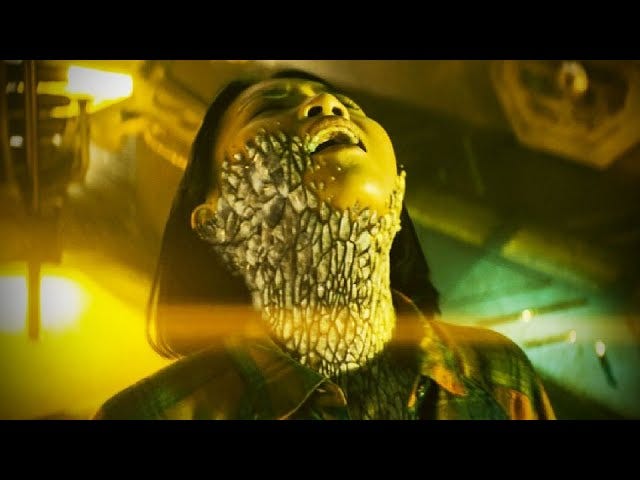 This image shows Suki Cheng (played by Molly Harris) tilting her head skyward and screaming in pain as the Praxeus bacterium overtakes her upper body. Large, tannish, stony encrustations cover Suki's neck and the right side of her face. This image shows Suki Cheng (played by Molly Harris) tilting her head skyward and screaming in pain as the Praxeus bacterium overtakes her upper body. Large, tannish, stony encrustations cover Suki's neck and the right side of her face.