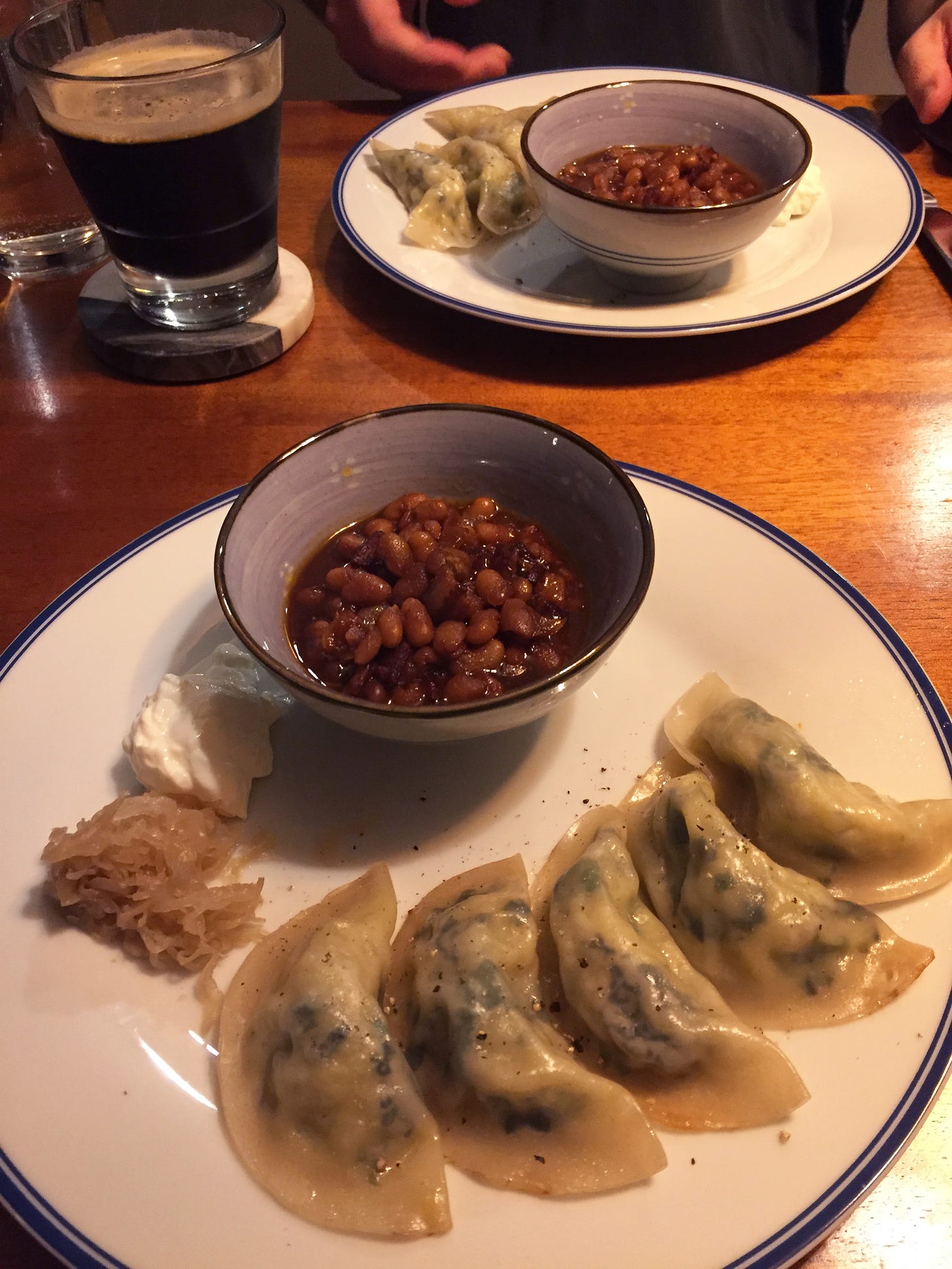 Two large plates with an arrangement of 5 pierogis each and a small amount of sour cream and sauerkraut, with a small purple bowl filled with baked beans. A glass of dark beer sits on a coaster next to the plate in the background.