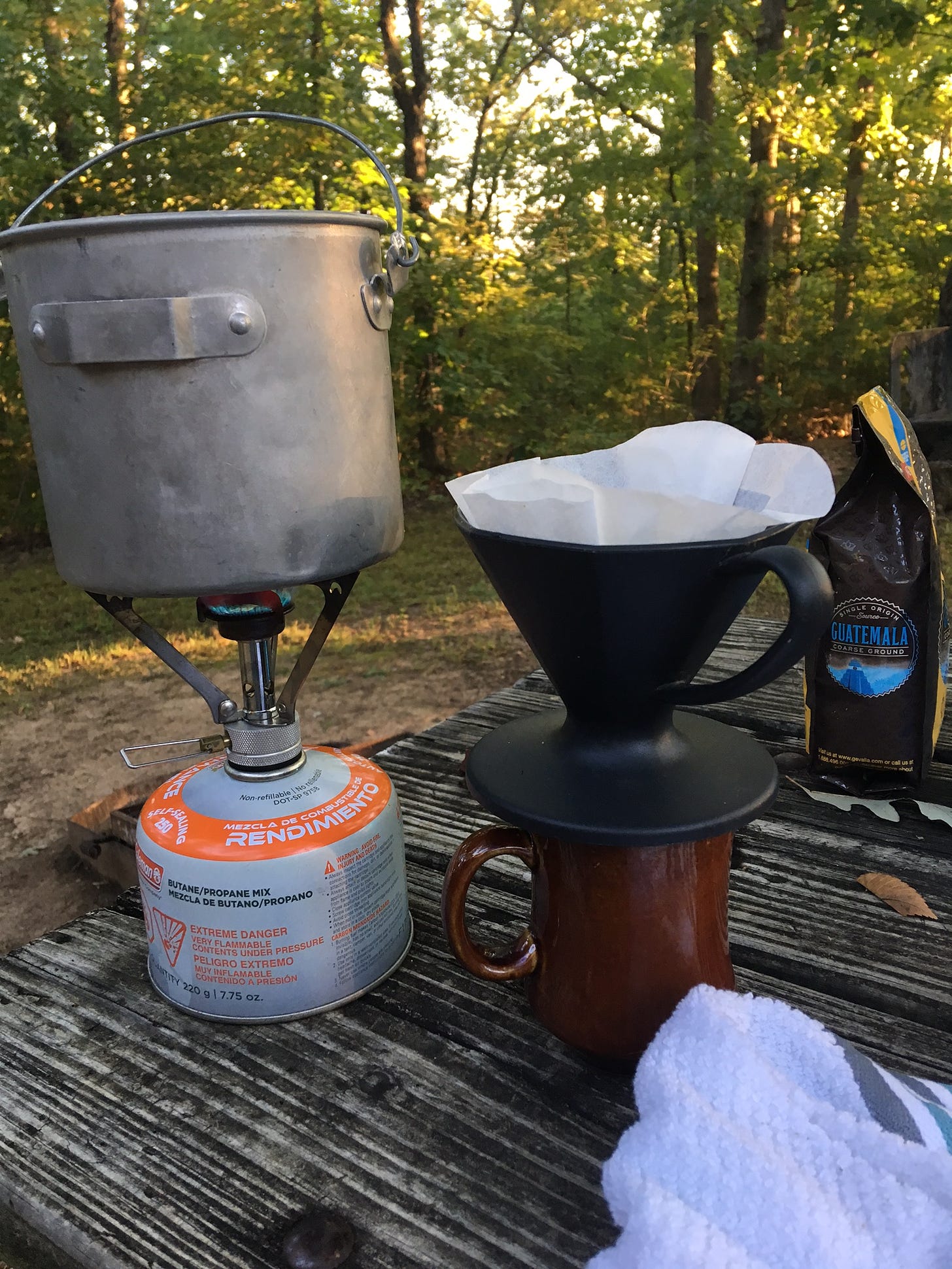 A pot on a small camping stove and a cup of coffee with brightly lit trees beyond