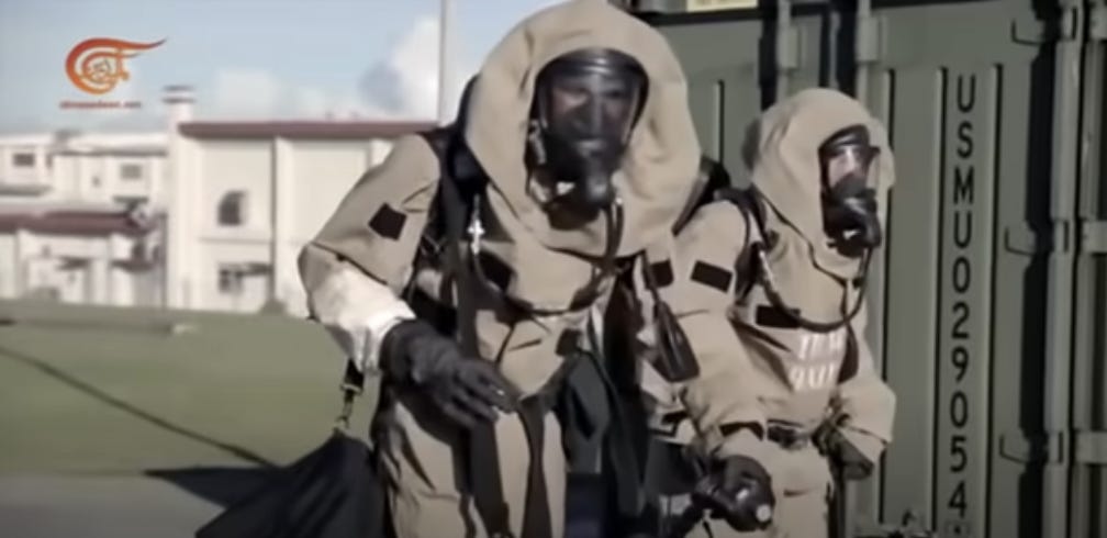 Picture of an outside scene with a building in the background. Two people are in the foreground wearing grey hazmat suits so only their eyes are visible behind blurry gas masks. They are walking next to a metal door or building that has a code written on it. What can be seen of the code reads vertically down the side: “USMU029054”.