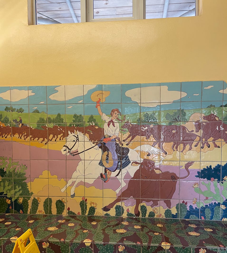 A picture of a rest stop bathroom in South Texas. The picture shows tile art of a white cowgirl on a white steed. Behind her, there's a herd of brown cattle, with a cowboy in the distance. There's hills and a blue sky. In the foreground, cacti bloom. The bottom half of the art is actuall a bench. The rest of the wall is beige, and a small window near the ceiling reveals wooden roofing outside.