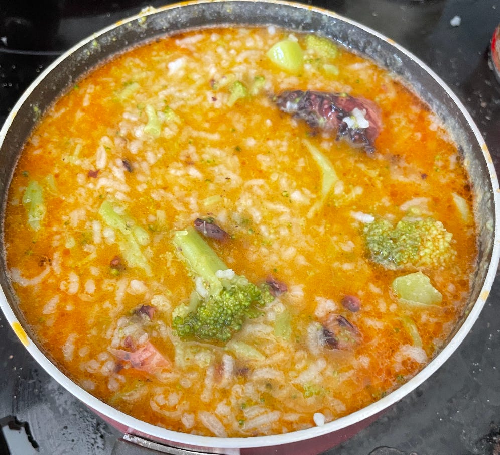 An image of a pot of congee (a rice porridge dish). Orange in color with broccoli and squid visible.