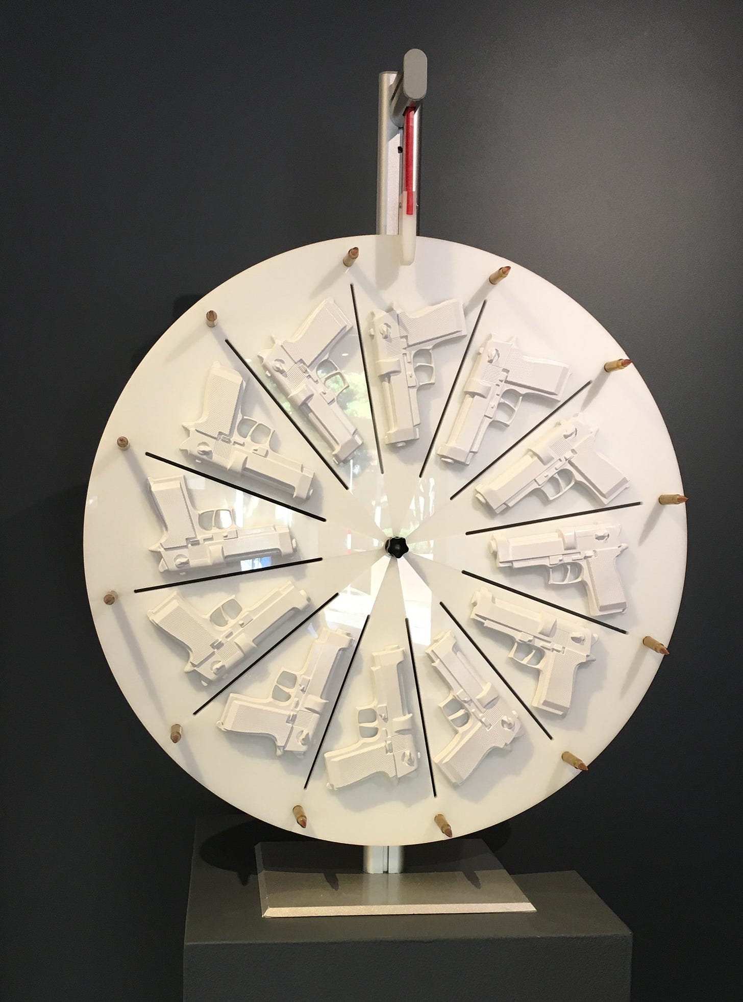 A white lacquered wheel, reminiscent of the Price is Right, is covered with twelve white handguns. At the top of the wheel is a flapper that hits the twelve shiny, gold bullets that stick out from the wheel when it is spun. 