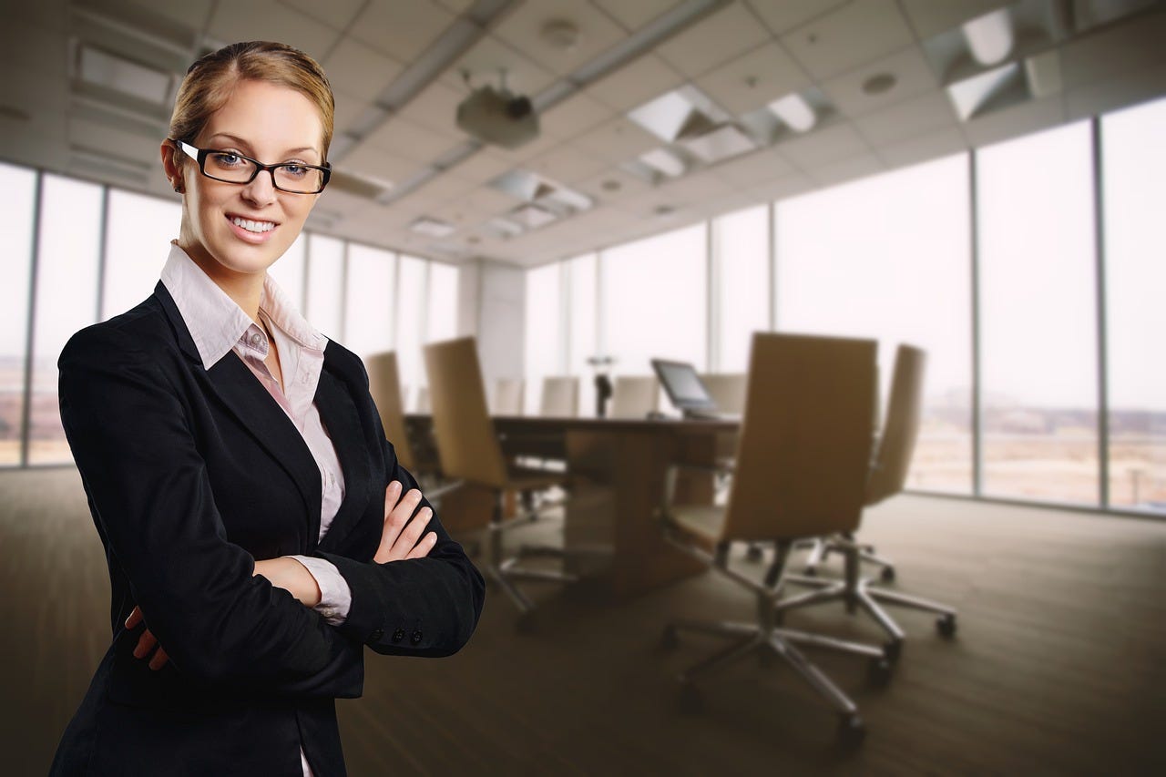 A white woman with blonde hair and glasses wearing a suit looks at the camera with her arms crossed in the foreground. In the background, a conference room