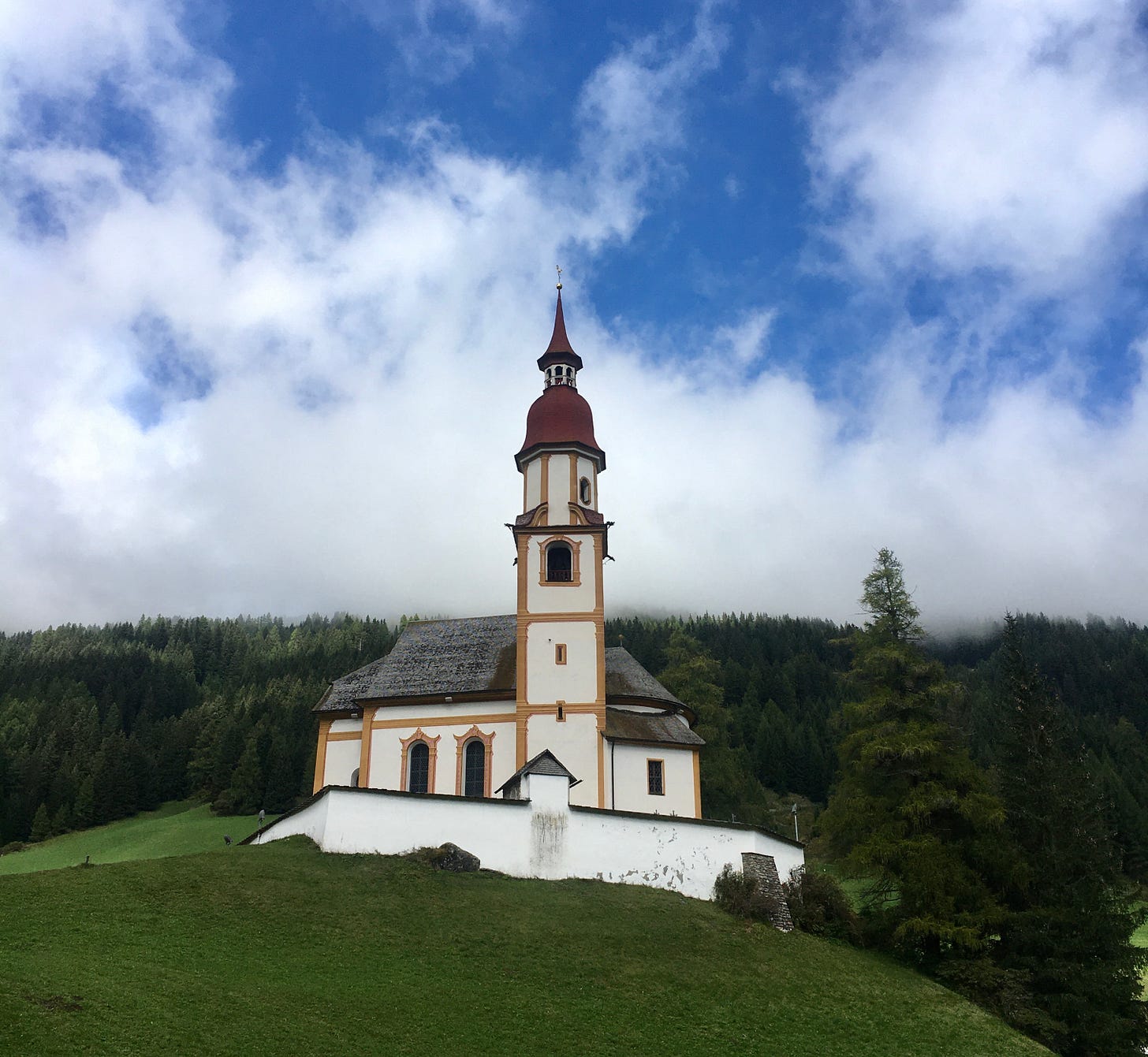 Eine Kapelle mit rotem Turm-Dach auf einem Hügel vor Wald, der Himmel ist blau mit Wolken