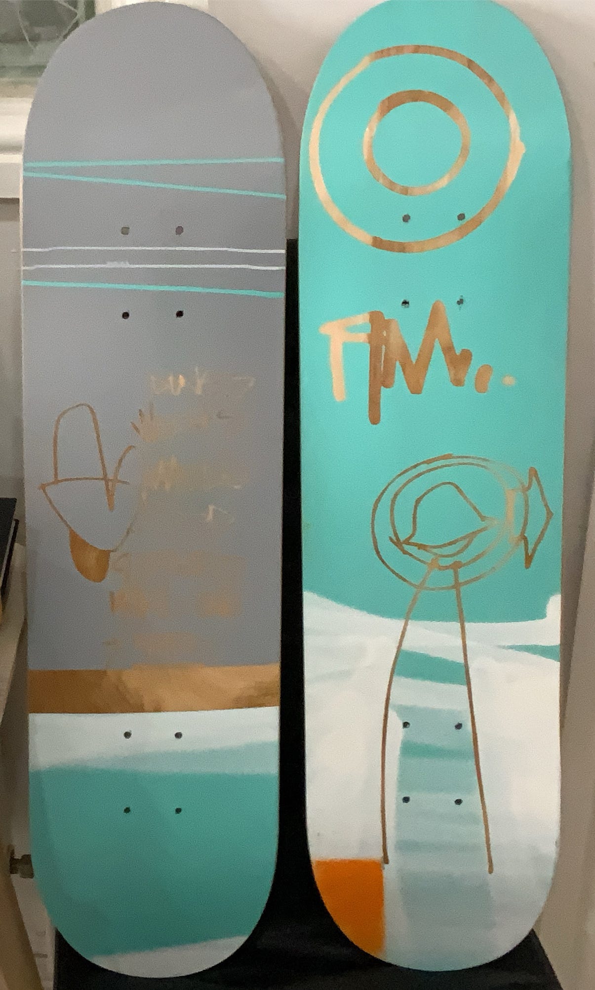 A photo of two painted skateboards without wheels. Various shades of teal, grey, white and gold intersected with Mylo Elliott's iconic grafitti handwriting.
