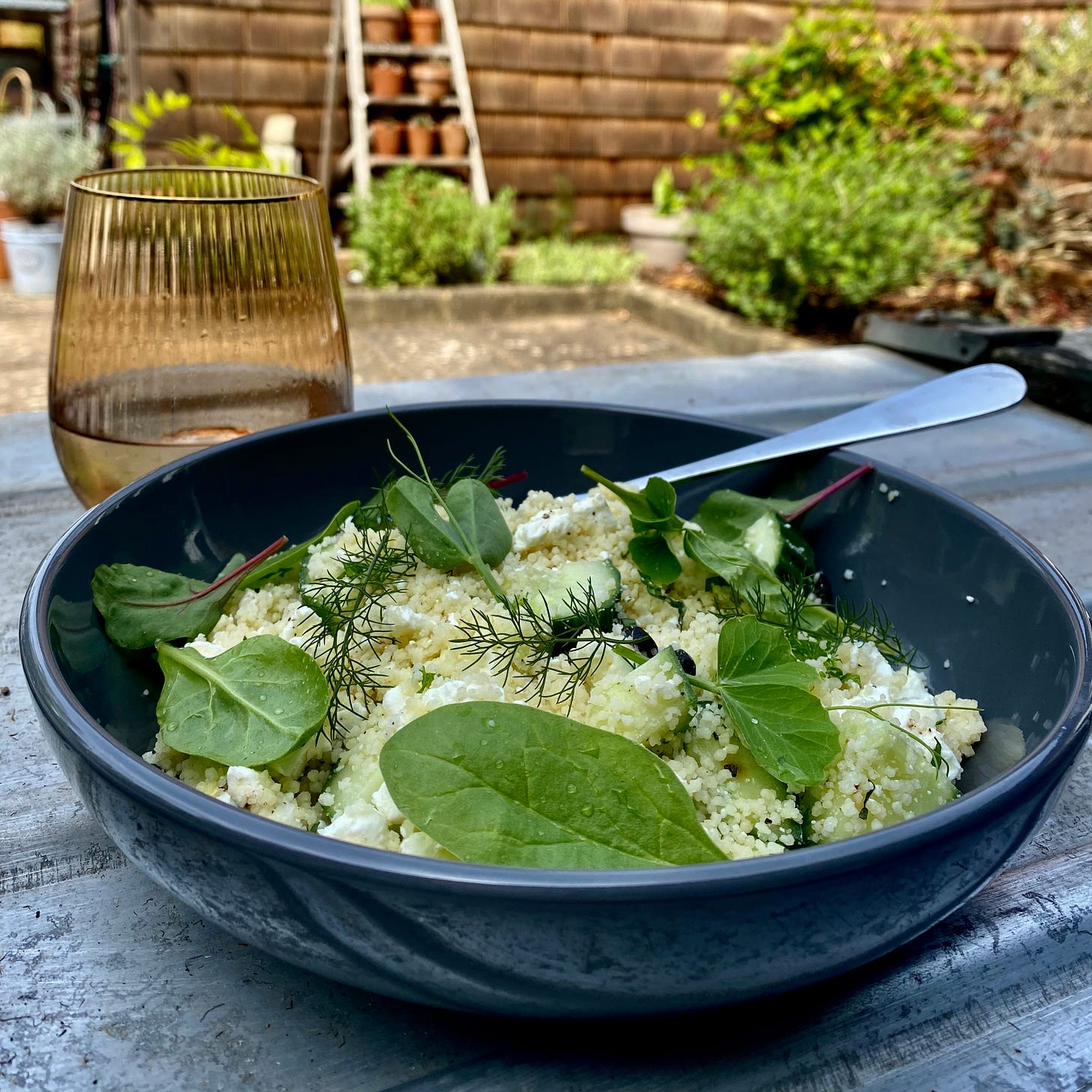 Bowl of couscous salad with feta and mint, scattered with salad leaves and herbs. Bowl is on a metal surface in the garden with a glass to the side and a fork in the bowl
