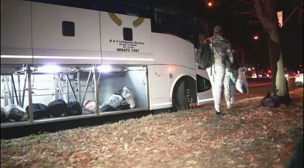 A person holding a plastic bag of belongings next to a bus with a luggage compartment visible at night.