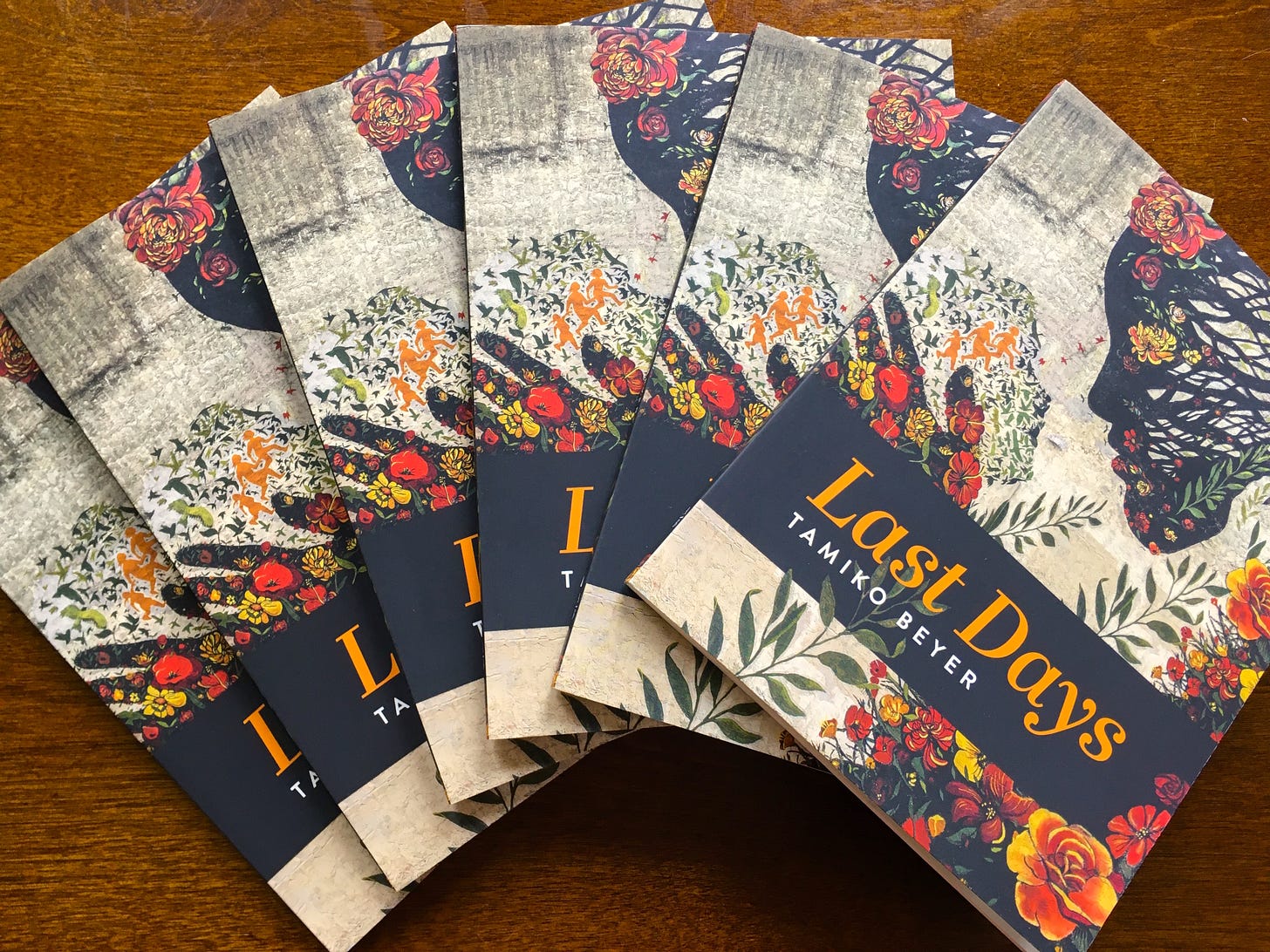 Five copies of Last Days, fanned out on a wooden table. The cover features two faces looking at each other. The faces are composed of branches, flowers, human figures, and birds. Text: Last Days, Tamiko Beyer
