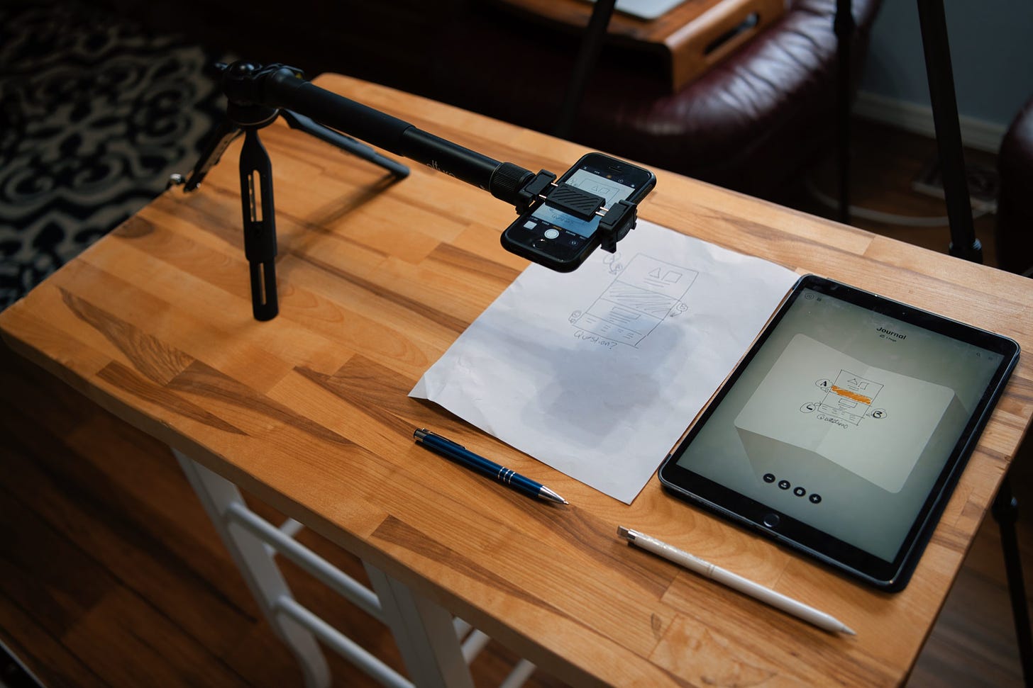 From an angle: Overhead shot of tripod with extnesion holding an iPhone with the video camera app open, paper and pen below it, iPad pro and pencil beside it