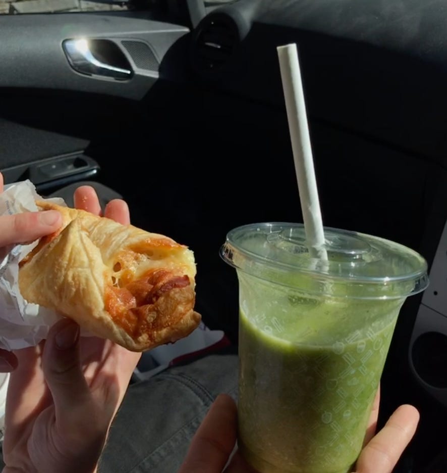 A photo taken from the driver’s seat of a van. Two white hands hold two different items up to the camera: a cheesy roll from Greggs in a white paper packet, to the left, and a bright green juice in a plastic cup with a straw. 