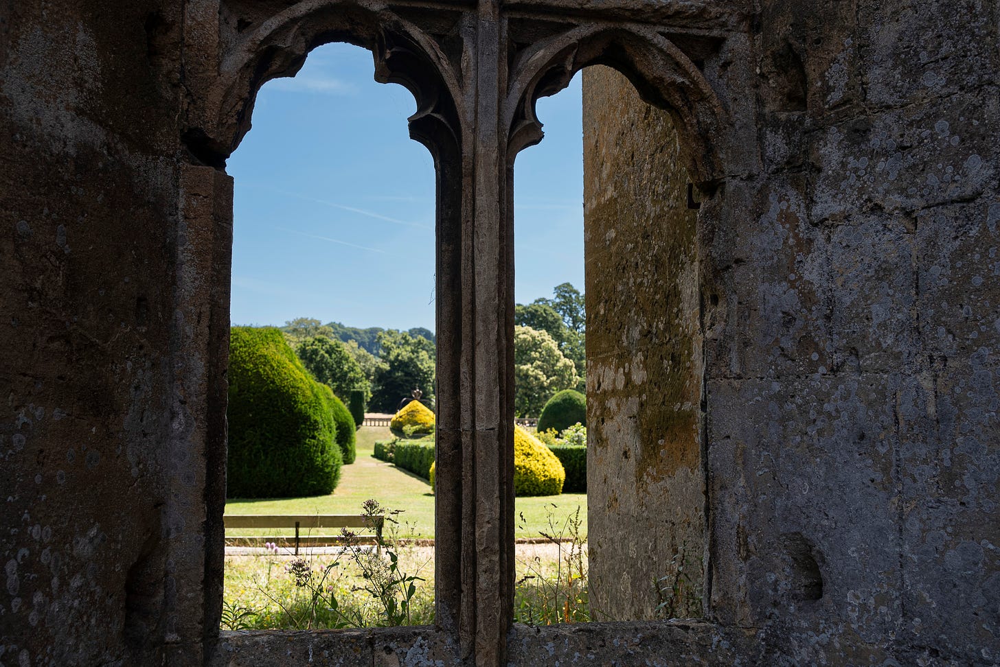A view through the window of a castle ruin looking out at the lush landscaped garden and a wooden bench
