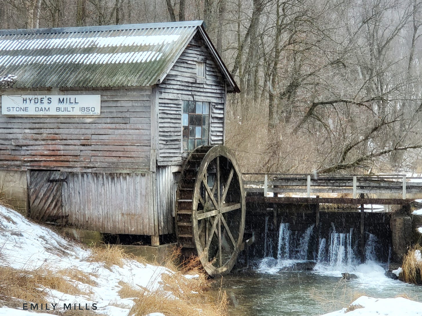 An old wooden building with a waterwheel borders a leaking stone dam and a low creek. Snow covers the brown grass on the shore below and snow can be seen falling in the air.