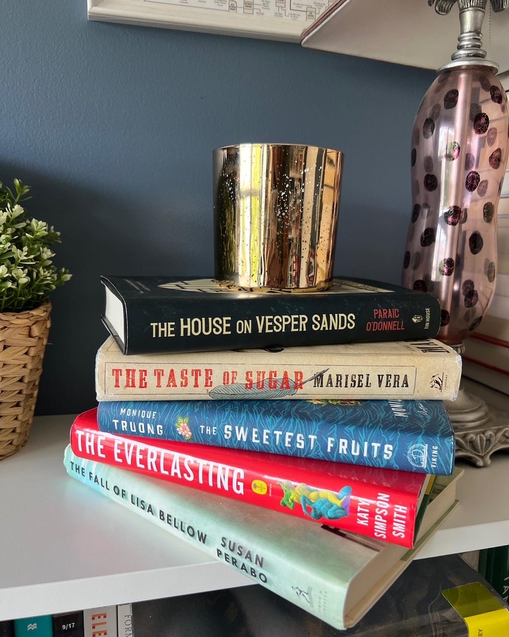 A stack of books with a candle on top. The books include The Fall of Lisa Bellow, The Everlasting, The Sweetest Fruits, The Taste of Sugar, and The House on Vesper Sands. 