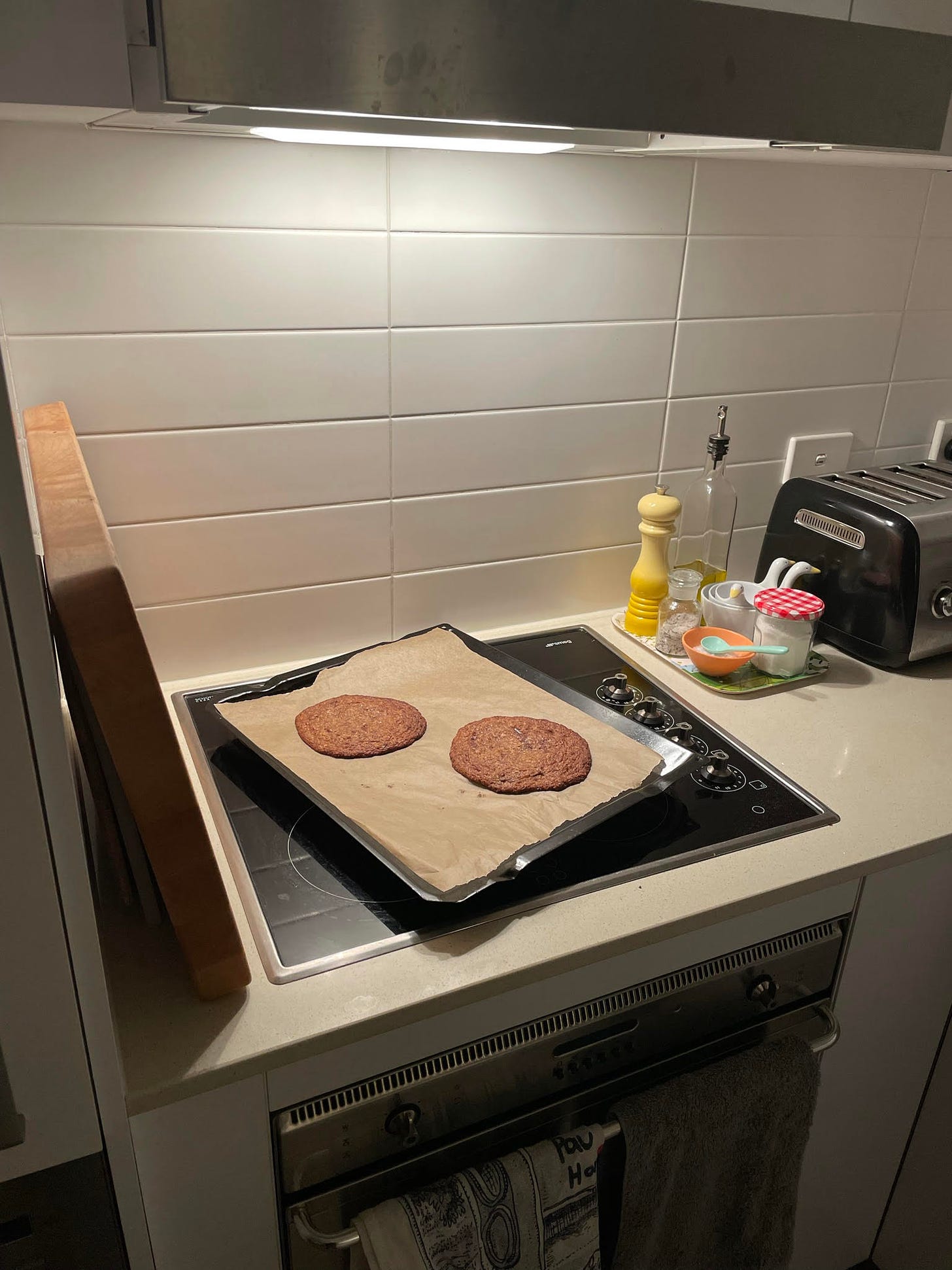 Two giant choc chip cookies on a baking tray, cooling on a stove at night.