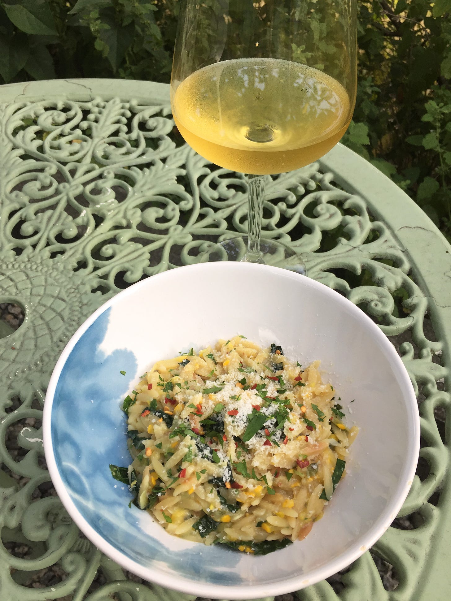 On a green wrought-iron table, a bowl of orzo with bits of kale and yellow zucchini visible throughout. On top is a sprinkling of parmesan, parsley, and chili flakes. A stemmed glass of white wine is behind the bowl.