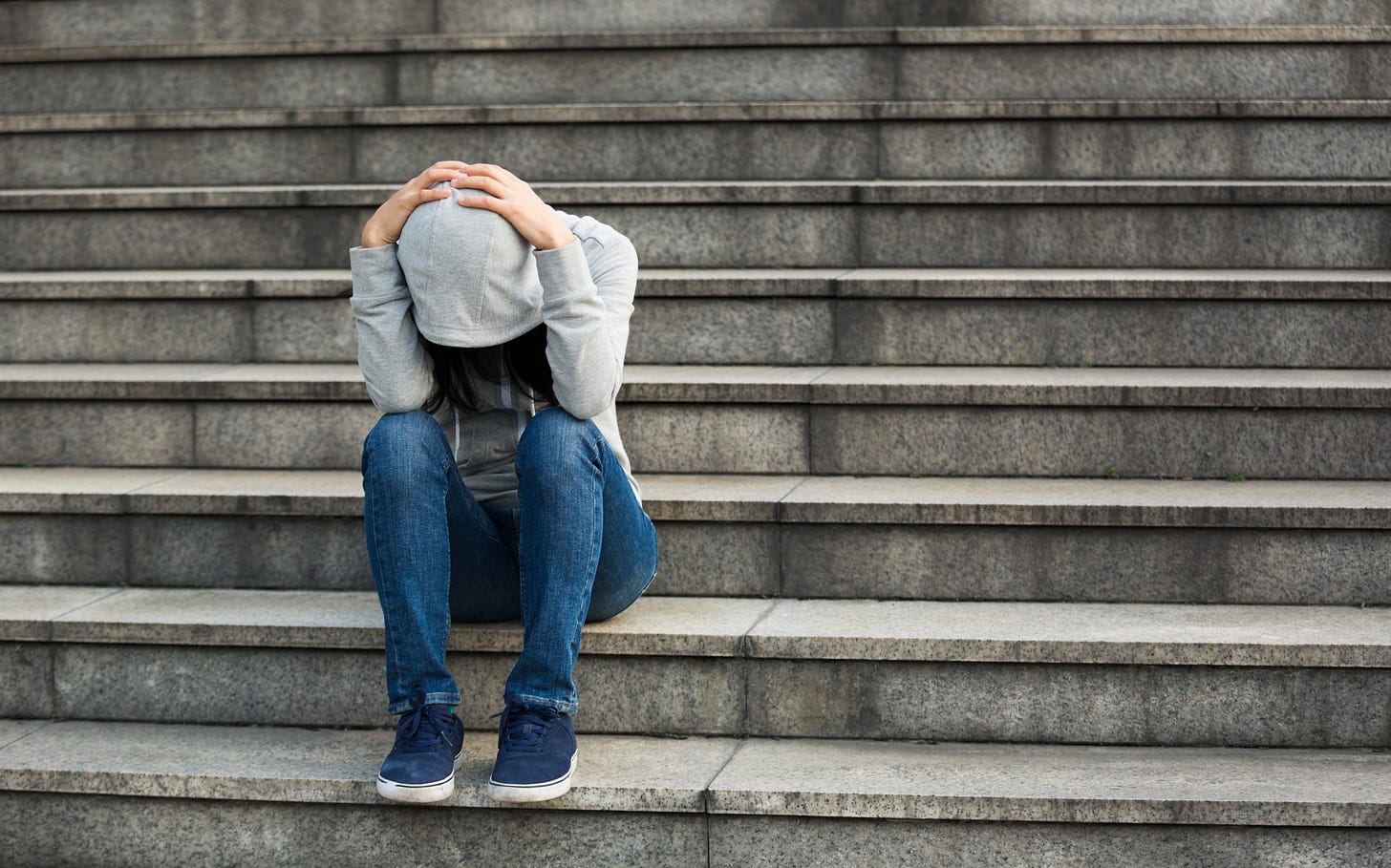A woman sits sadly on stairs with her head in her hands and her hood pulled up.
