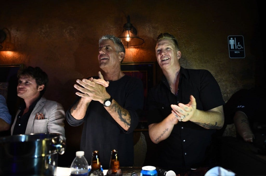 Anthony Bourdain and Queens of the Stone Age frontman Josh Homme stand next to each other in a dimly-lit room. They are both clapping.