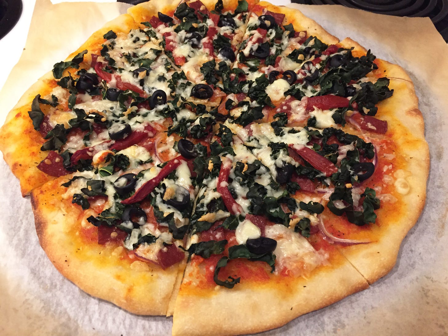 On parchment paper atop a pizza stone, a kale and olive pizza with browned edges and slices of roasted red pepper visible amid the cheese.
