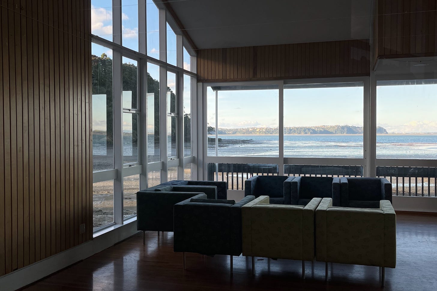 A photograph looking out of a striking set of midcentury windows out to a low-tide bay in the Manukau Harbour. In the foreground is a sectional set of corporate-looking armchairs, and the walls are panelled with timber. The room is dark. The hour is late in the day, with low-angled blue light on the bay.