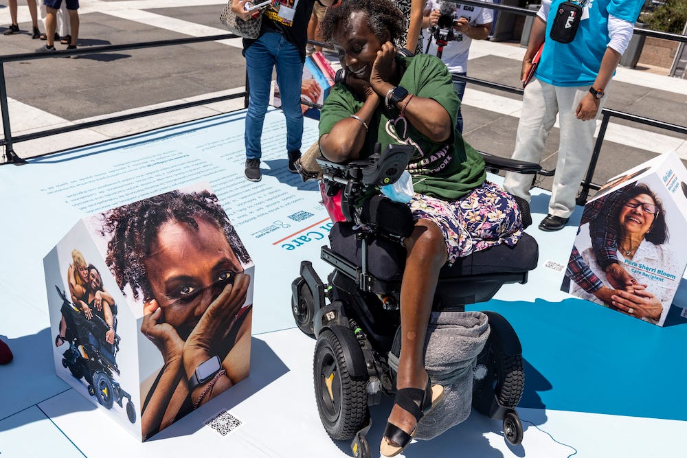 photo of Engracia looking at square sculpture of herself, while sitting in wheelchair, with green hand in hand shirt. While she holds her head in the palm of her hands with a shocked expression.