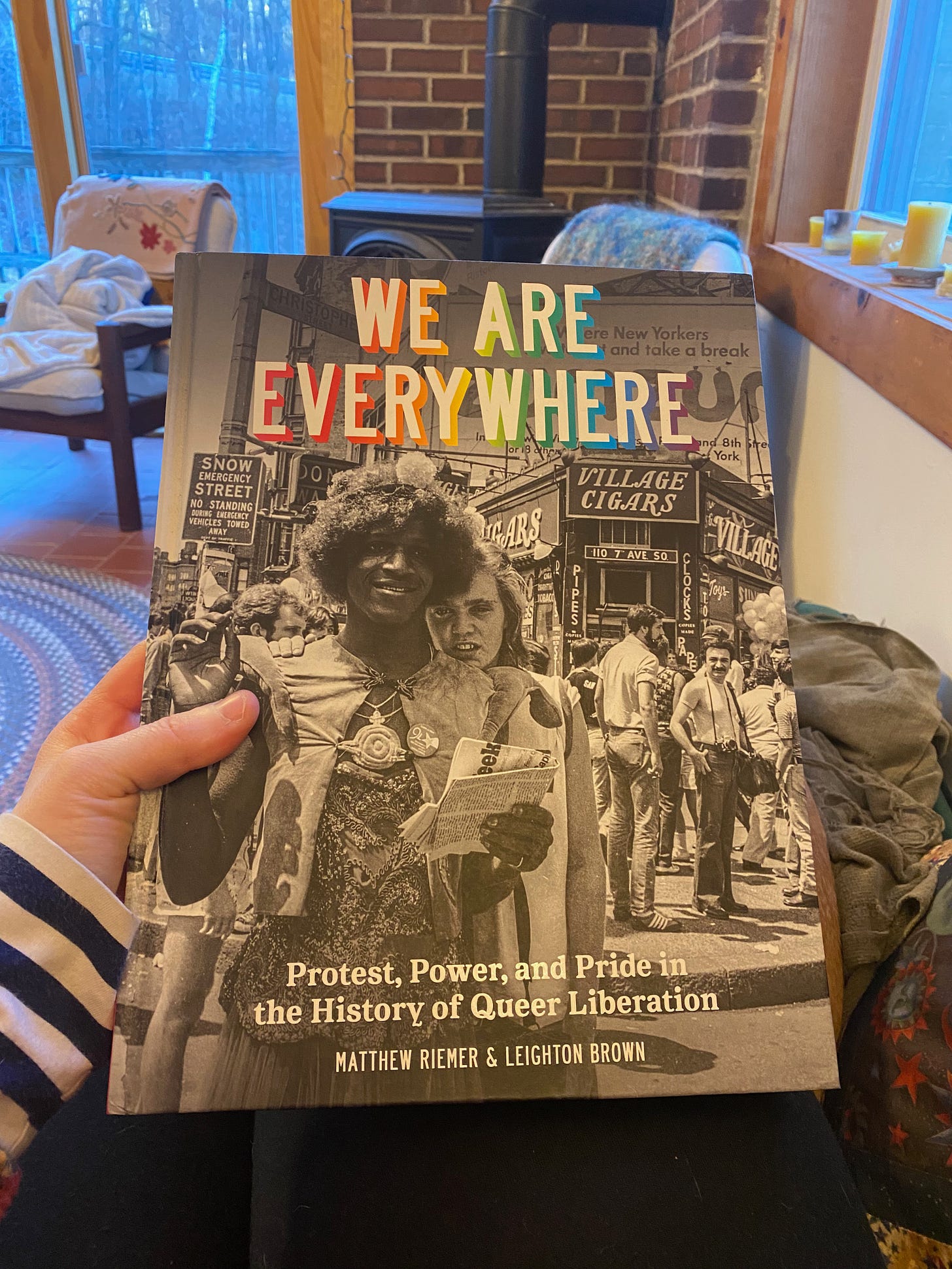 My hand holding up the photo book We Are Everywhere by Matthew Riemer and Leighton Brown. It is a large hardcover with a black and white photograph of Marsha P. Johnson on the cover. An armchair and windows looking out into the woods are visible in the background.