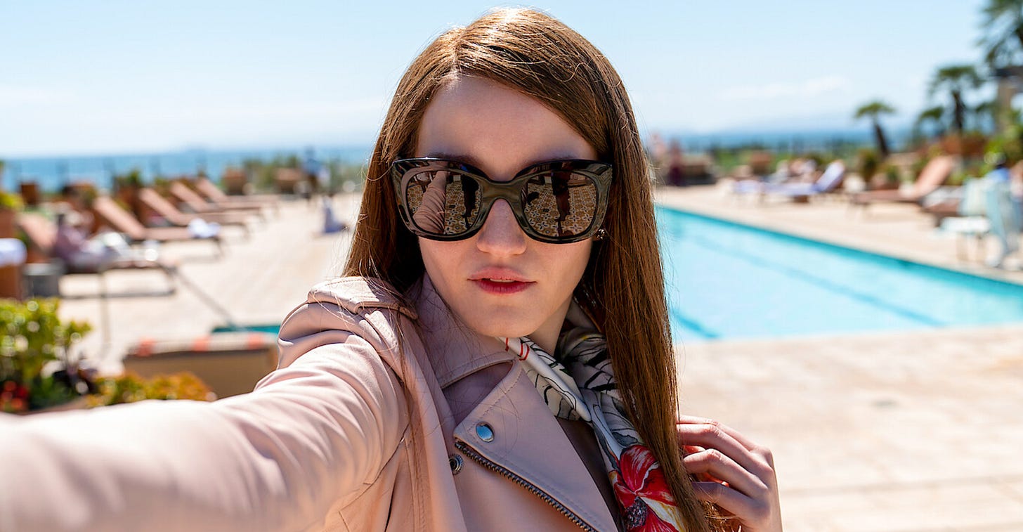 An image of a brunette woman standing infront of a pool, wearing large sunglasses, with patterned tile in the reflection of the sunglasses.  The woman is the actress Julia Garner, portraying the role of Anna Delvey, from the Netflix series "Inventing Anna."