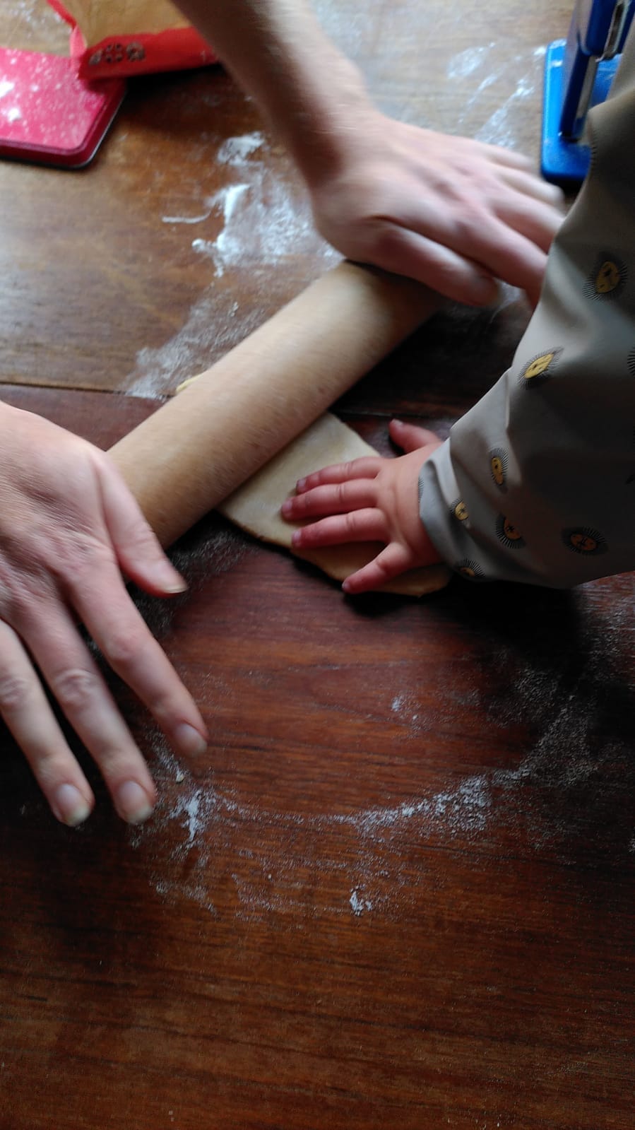 An action shot of Kate's white hands putting pressure on a rolling pin, while a toddler's little white hands are touching the yellow dough, helping out. The work surface is a wooden table with white flour dusted haphazardly. 
