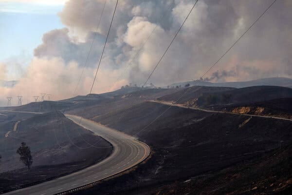 A wide view of hills blackened from fire, with an empty highway cutting through. Giant clouds of smoke billow into the sky on the horizon, with transmission lines leading to electricity pylons in the distance.