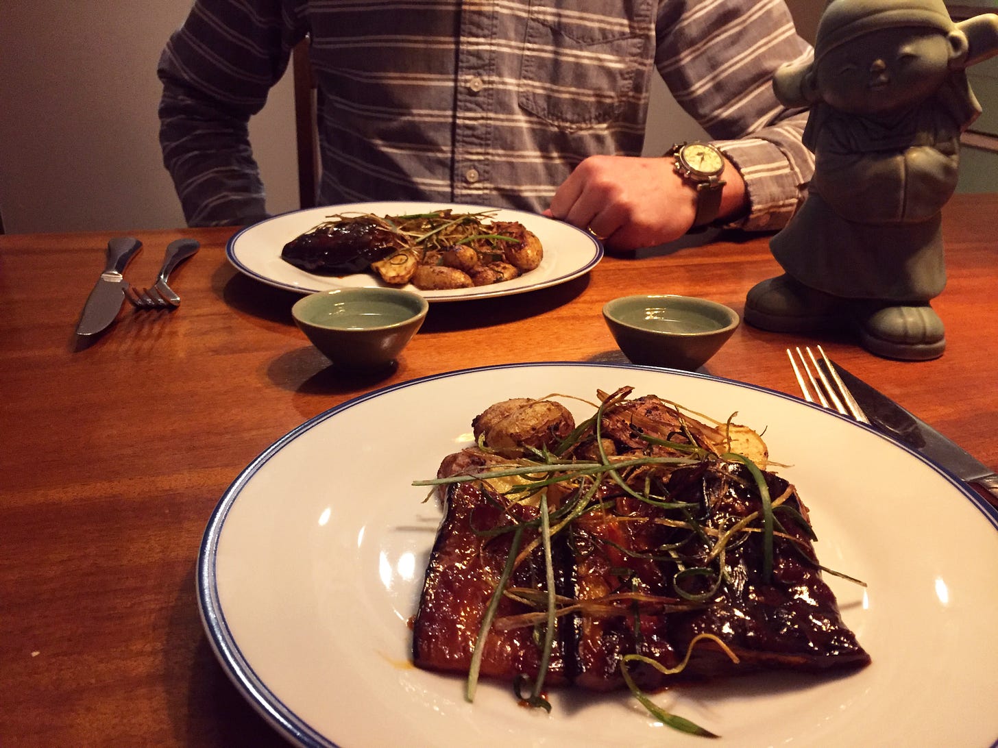 Two plates with large slices of eggplant covered in a dark red, sticky glaze, arranged overlapping a pile of small fingerling potatoes. On top are long thin slices of crisp green onion. Two mint green cups of soju sit between the plates, and just visible in the right corner is a matching ceramic bottle with a human shape.