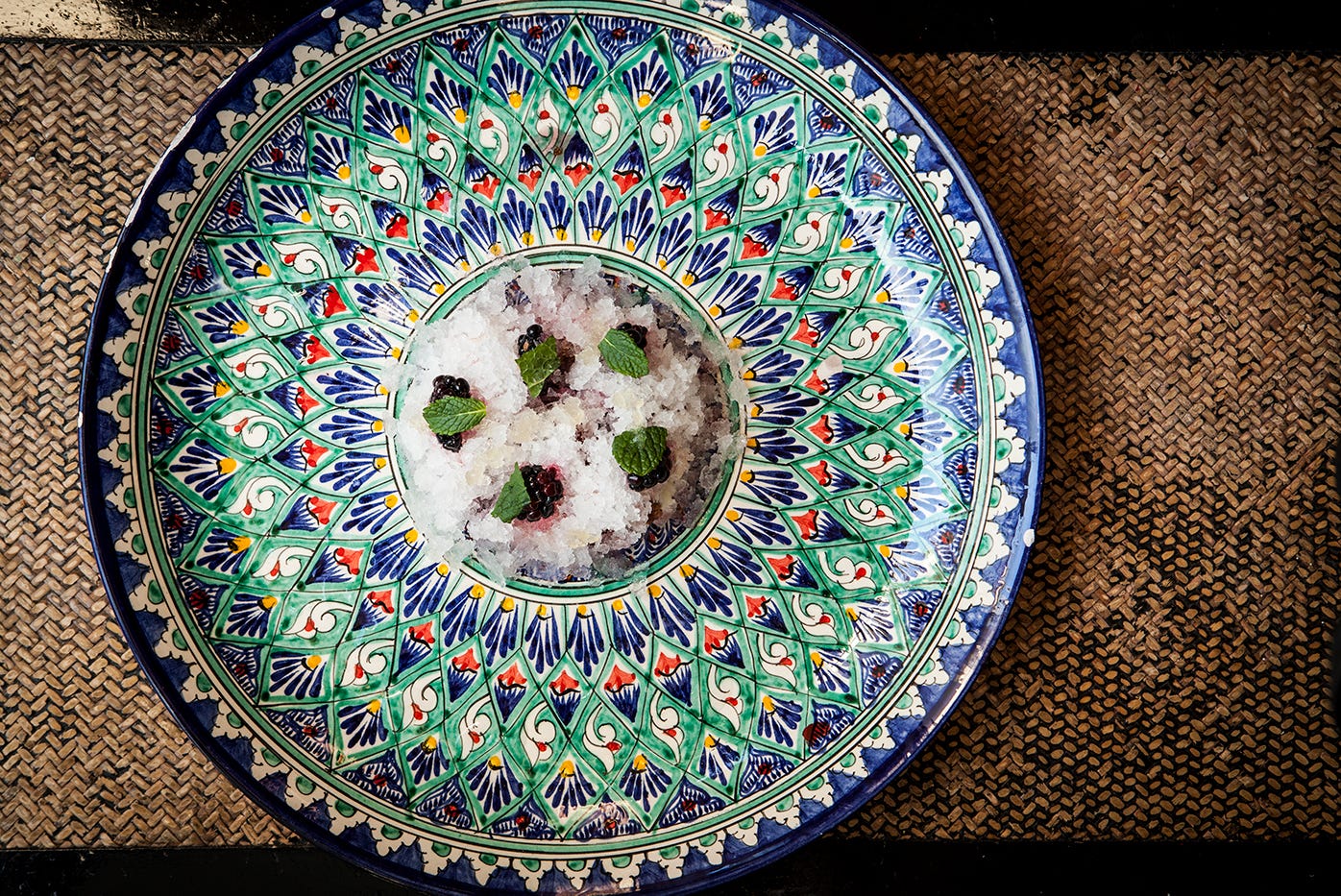 An overhead of photo of a large colourful plate on a rattan mat. In the centre of the plate is a pile of granita, decorated with black mulberries, mint leaves and pieces of mochi.
