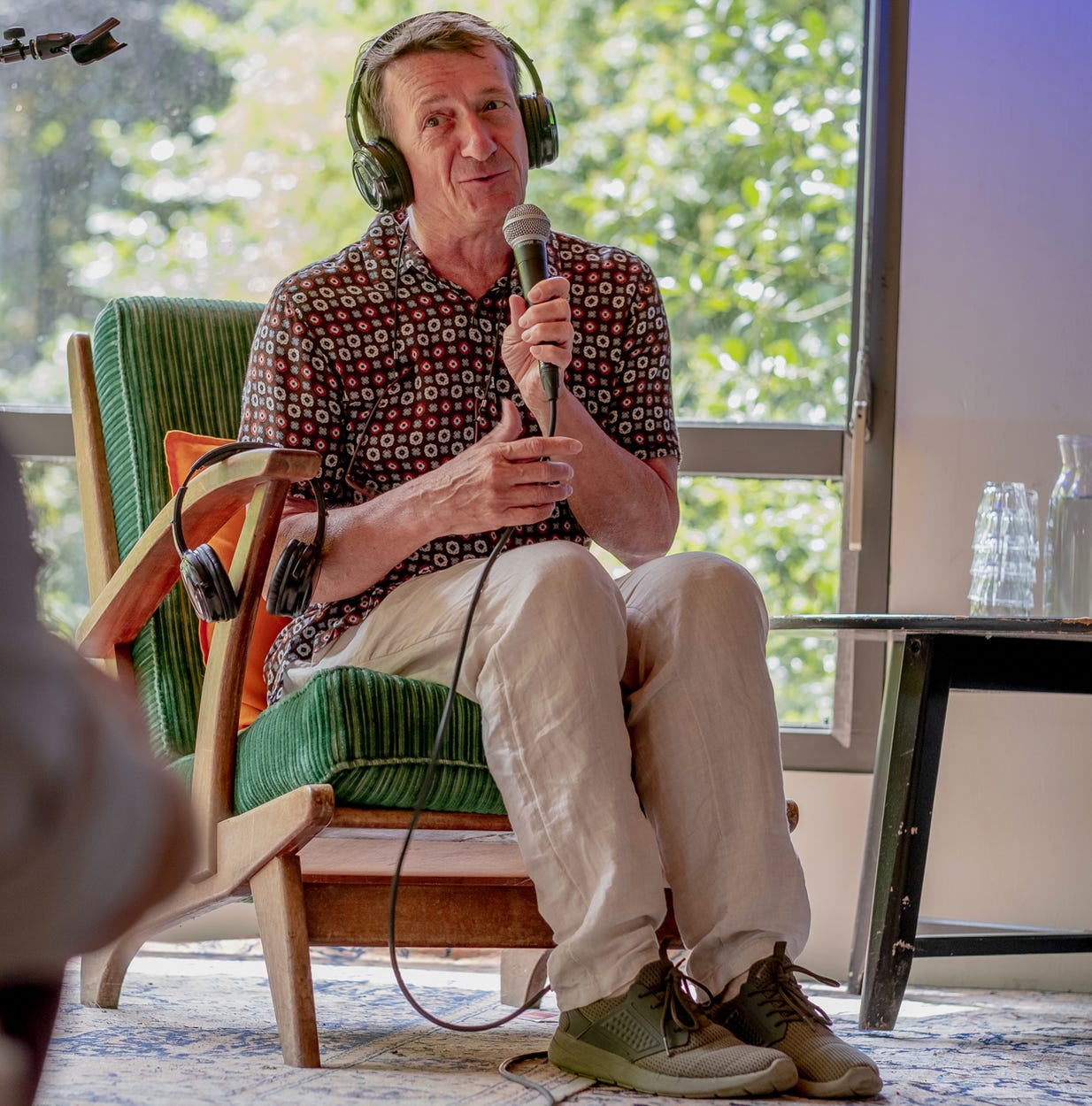 foto van een oudere man die op een podium op een stoel zit. Hij draagt een beige broek en een bloemetjesshirt. In zijn hand een microfoon, op zijn hoofd een koptelefoon