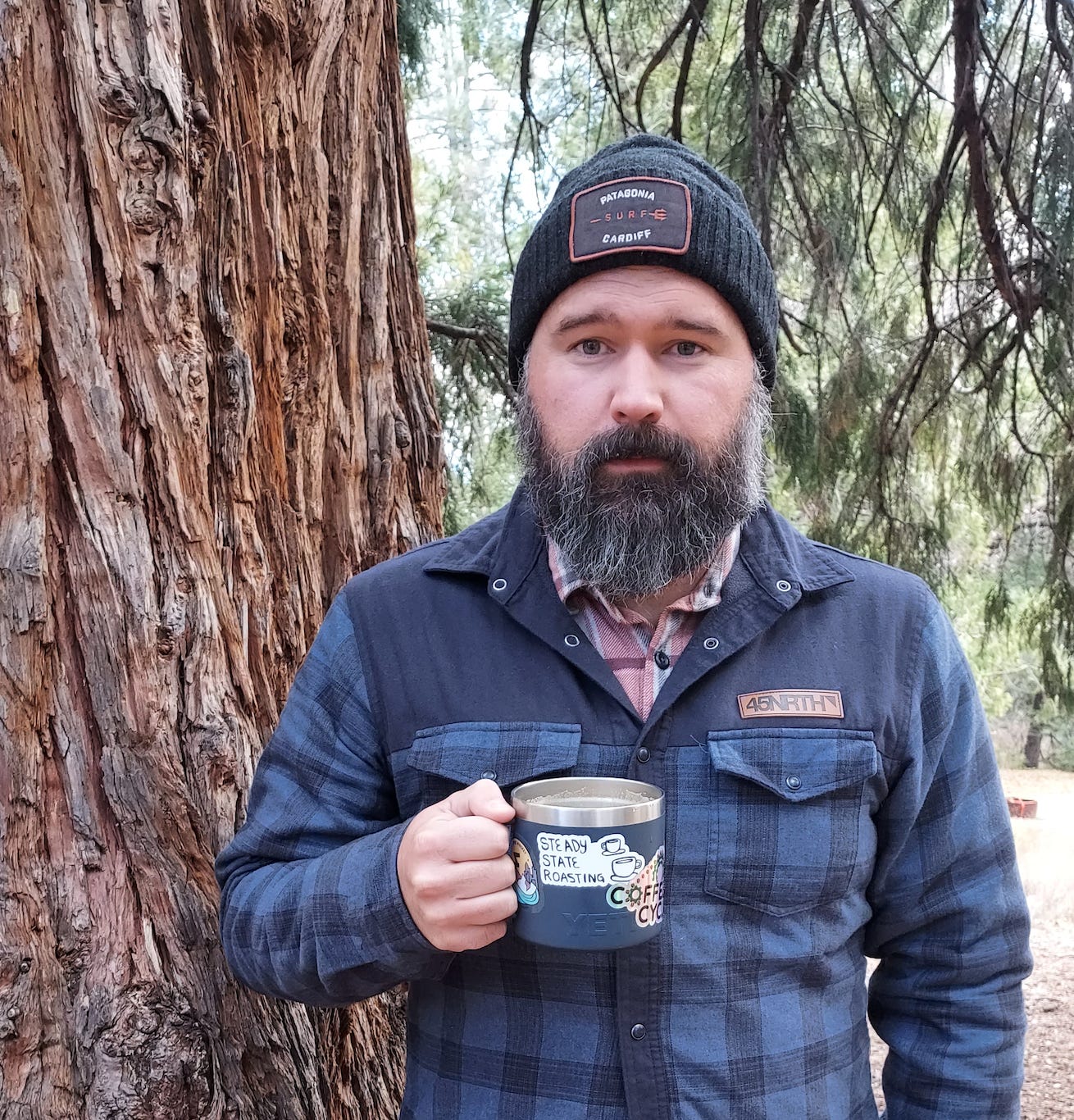 A white guy with a salt and pepper beard wearing a grey stocking cap and dark blue flannel coat holds a navy Yeti stainless steel coffee mug and leans up against a California Pine Tree.